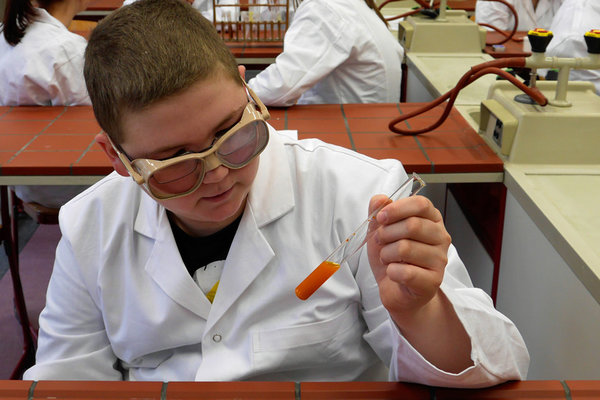 Ein Schüler mit Schutzbrille und Laborkittel schaut sich ein Reagenzglas mit orangenem Inhalt an, welches er in der Hand hält.