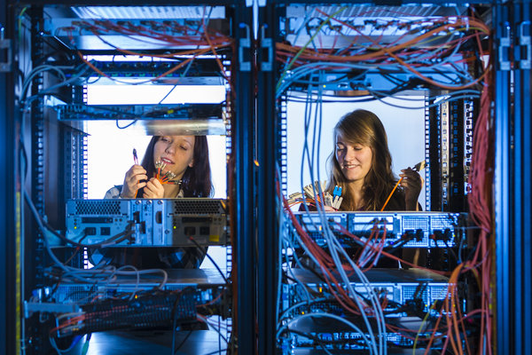 Blick durch technische Geräte auf zwei Auszubildende. Die beiden Mädchen überprüfen Kabel und Steckverbindungen.