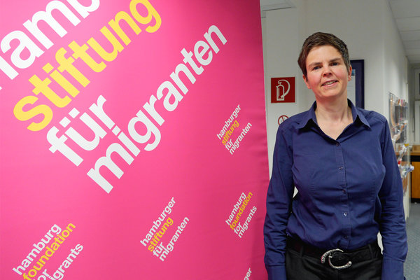 Susanne Dorn steht vor einer pinken Wand auf der "Hamburger Stiftung für Migranten" steht.