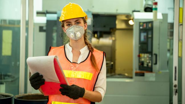 Eine Frau mit Maske, Bauhelm und Warnweste schaut in einer Werkhalle direkt in die Kamera